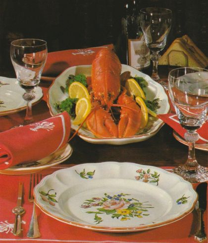 Ein ganzer gekochter Hummer auf einem ovalen Teller, garniert mit Zitronenscheiben und Petersilie, steht im Zentrum einer festlich gedeckten Tafel. Das Gedeck umfasst orange Stoffservietten mit Hummermotiv, Weingläser mit geschliffenem Stiel und Porzellangeschirr mit Blumenmuster.