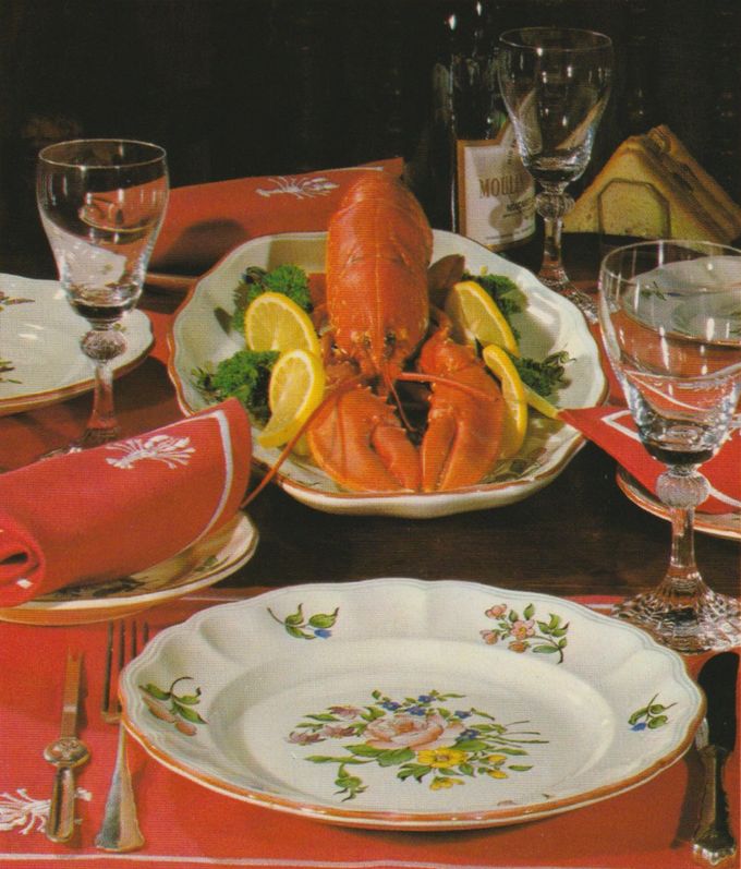 Ein ganzer gekochter Hummer auf einem ovalen Teller, garniert mit Zitronenscheiben und Petersilie, steht im Zentrum einer festlich gedeckten Tafel. Das Gedeck umfasst orange Stoffservietten mit Hummermotiv, Weingläser mit geschliffenem Stiel und Porzellangeschirr mit Blumenmuster.