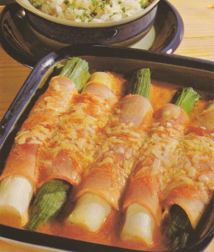 In einer dunklen, quadratischen Auflaufform: mit Schinken umwickelte Porreestangen in Tomatensoße, überbacken mit goldbraun geschmolzenem Käse. Die grünen Porree-Enden ragen an beiden Seiten heraus. Im Hintergrund eine dunkle Schüssel, vermutlich mit Reis als Beilage.