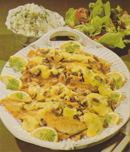 Gebratene Seezungen auf einer ovalen Servierplatte, gefüllt mit Champignons in Sahnesauce, garniert mit Nordseekrabben, Petersilie und Zitronenscheiben. Im Hintergrund eine Schale mit Petersilienreis und ein Teller mit Kopfsalat–Tomaten–Salat.