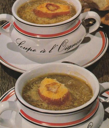 Zwei Suppentassen mit klarer Zwiebelsuppe und überbackener Käsekruste stehen auf einer weißen Servierplatte mit orangefarbenem Rand und der Aufschrift „Soupe à l’Oignon“. Daneben liegen Suppenlöffel auf dem dunklen Holztisch.