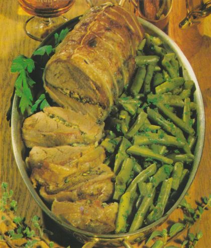 Aufgeschnittene Schweinefilet-Rolle, mit Kräuter-Füllung und Räucherspeck-Ummantelung, auf einer ovalen Metallplatte. Daneben gebutterte Brechbohnen mit gehackter Petersilie. Garniert mit frischer Petersilie und Majoran-Zweigen.