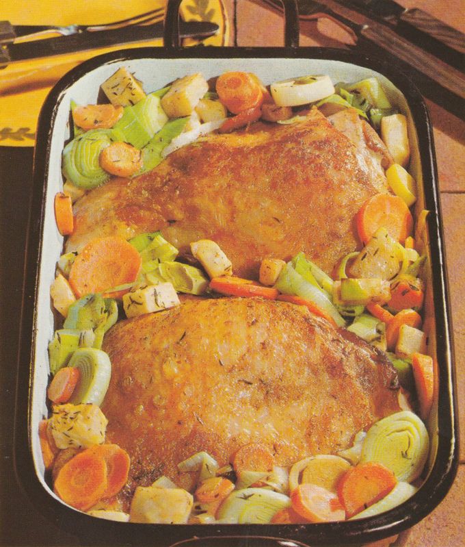 Zwei gebratene Puten–Oberkeulen in einer rechteckigen Bräterform, umgeben von geschmortem Gemüse: Porree–Ringe, Möhren–Scheiben, Sellerie– und Knoblauch–Stücke. Die Keulen haben eine knusprige Haut.