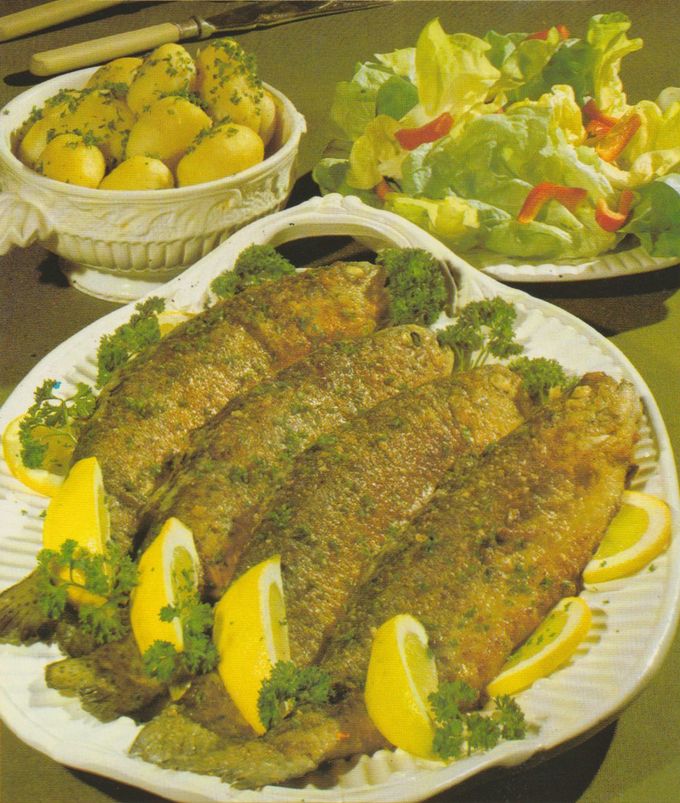 Knusprig gebratene Forellen auf einer ovalen Platte, garniert mit Zitronenscheiben und Petersilie. Im Hintergrund Pellkartoffeln mit gehackter Petersilie in einer Schüssel und ein gemischter Blattsalat mit Paprikastreifen.