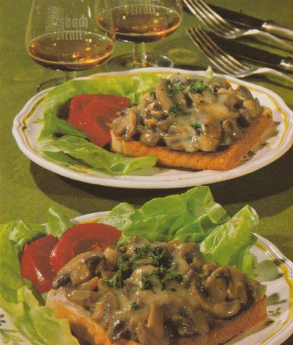 Panierte Toastscheiben mit Champignon–Rahm–Sauce, garniert mit Petersilie, auf einem Bett aus Salatblättern mit Tomatenscheiben. Im Hintergrund zwei Gläser Asbach Uralt.