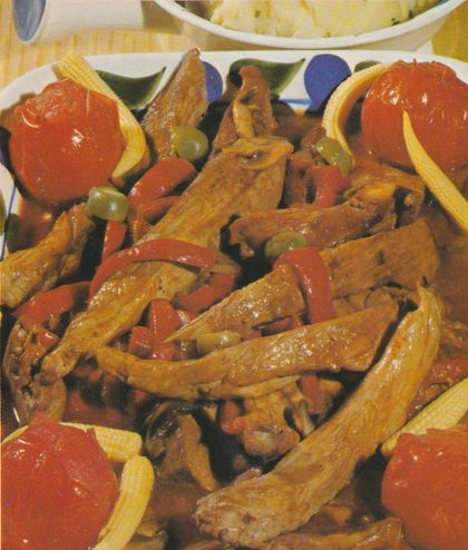 Gebratene Schweinefilet-Streifen in dunkler Sauce mit roten Paprika-Stücken, grünen gefüllten Oliven und Champignons auf einem blau-weiß gemusterten Teller. Garniert mit ganzen Schmortomaten und Maiskolben-Stücken. Im Hintergrund eine Schüssel mit Kartoffelpüree.