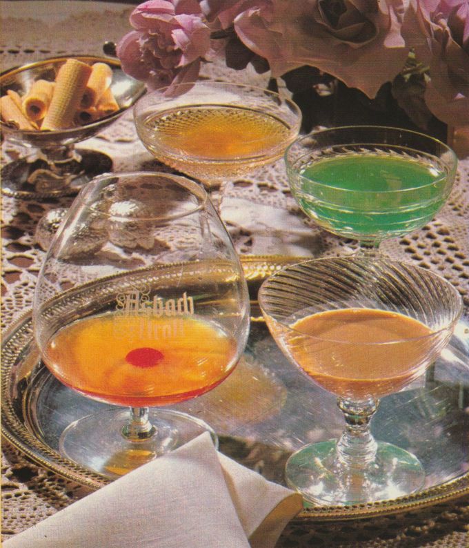 Vier verschiedene Cocktails und Aperitifs in eleganten Gläsern auf silbernen Tabletts, darunter eine grüne, eine gelbe und eine rosafarbene Kreation, dekoriert mit einer Cocktailkirsche. Im Hintergrund rosa Rosen als Dekoration und eine Schale mit aufgerollten Gebäckröllchen.