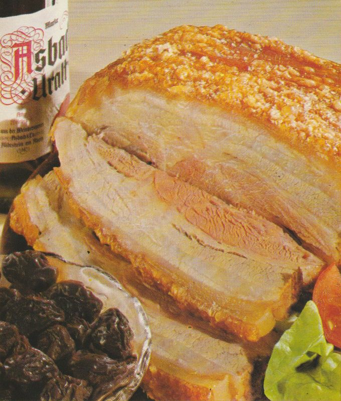 Aufgeschnittener Schweinebauch nach dänischer Art mit knuspriger, rautenförmig eingeschnittener Schwarte. Im Vordergrund eine Schale mit in Asbach-Uralt-Weinbrand marinierten Backpflaumen, links im Hintergrund eine Flasche „Asbach Uralt”.