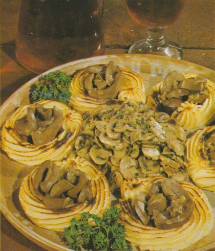 Auf einer großen, ovalen Keramikplatte angerichtete Kartoffelnester aus gesprittem, leicht gebräuntem Kartoffelpüree, gefüllt mit in Rotwein-Weinbrand-Sauce geschmorten Schweinenieren-Scheiben. In der Mitte der Platte cremige Champignons mit gehackter Petersilie. Frische Petersilienzweige als Dekoration. Im Hintergrund eine Flasche und ein Glas auf einer Holzfläche.