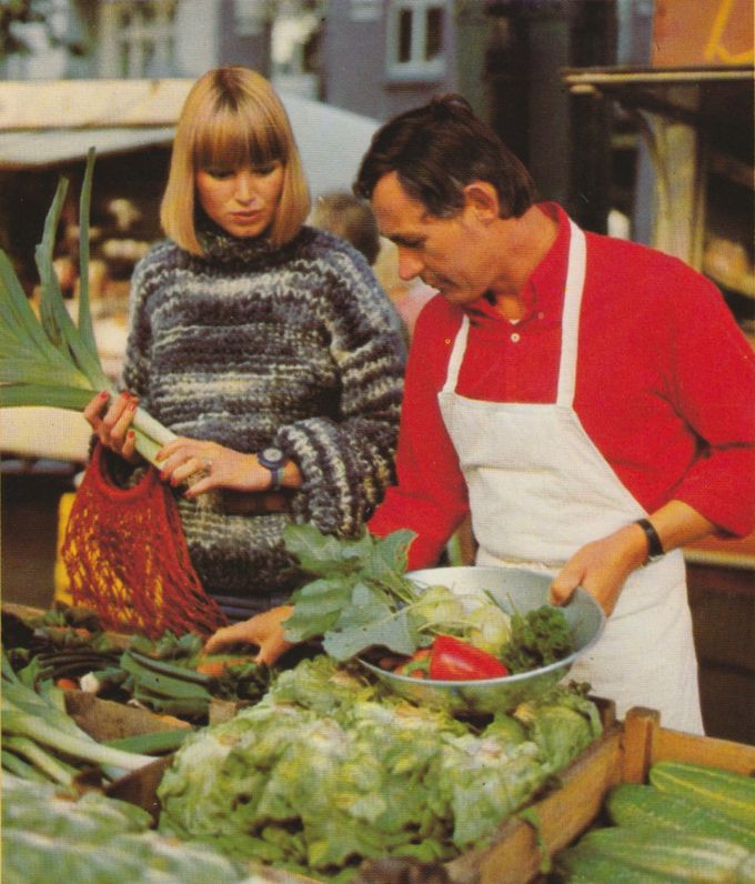 Eine Frau in gemustertem Pullover und ein Mann mit weißer Schürze betrachten eine Auslage mit frischem Gemüse, darunter Lauch, Salat, Bohnen und Tomaten.