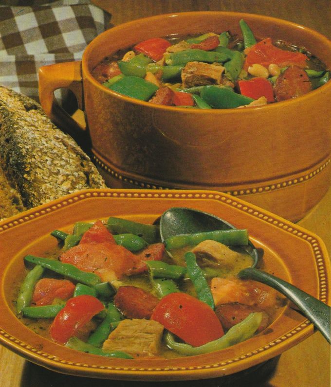 Herzhafter Eintopf mit Fleischstücken, grünen Bohnen, roten Paprikain orangefarbenen Schüsseln serviert. Daneben liegen Scheiben von dunklem Körnerbrot auf einem braun-weiß karierten Tischtuch.