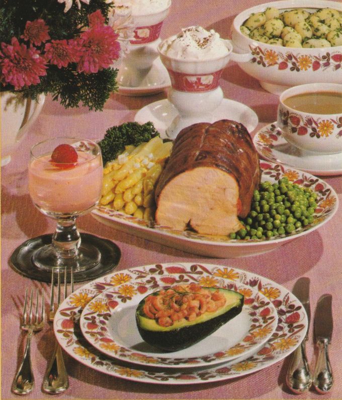 Ein festliches Menü mit Fleischbraten, Spargel und Erbsen auf einer Servierplatte. Auf dem Teller im Vordergrund liegt eine gefüllte Avocadohälfte mit Krabben. Dabei steht ein rosa Dessert im Glas. Die Tafel ist mit Blumenmustergeschirr, rosa Tischdecke, Kaffeetassen und einem Blumenstrauß dekoriert.