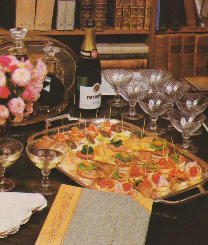 Eine große Servierplatte mit verschiedenen belegten Brotscheiben und Canapés, aufgespießt mit Zahnstochern, garniert mit Käse, Wurst, Tomaten und Petersilie. Im Hintergrund stehen Champagner- und Weinflaschen, geriffelte Kristallgläser, rosa Blumen und antike Bücher in einem Regal.