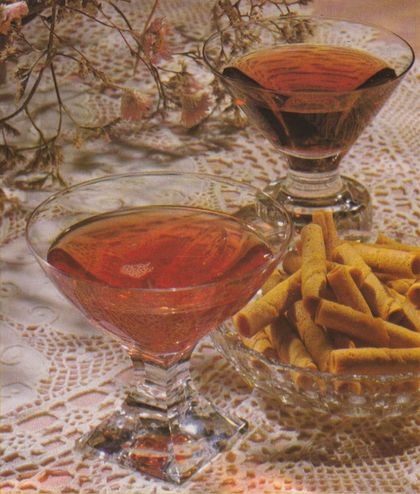 Zwei elegante Cocktailgläser mit bernsteinfarbener Flüssigkeit stehen auf einer gehäkelten Spitzendecke, daneben eine Glasschale mit goldbraunen gerollten Waffelröllchen. Getrocknete Blumen im Hintergrund runden die Dekoration im typischen Stil der 1970er-Jahre ab.