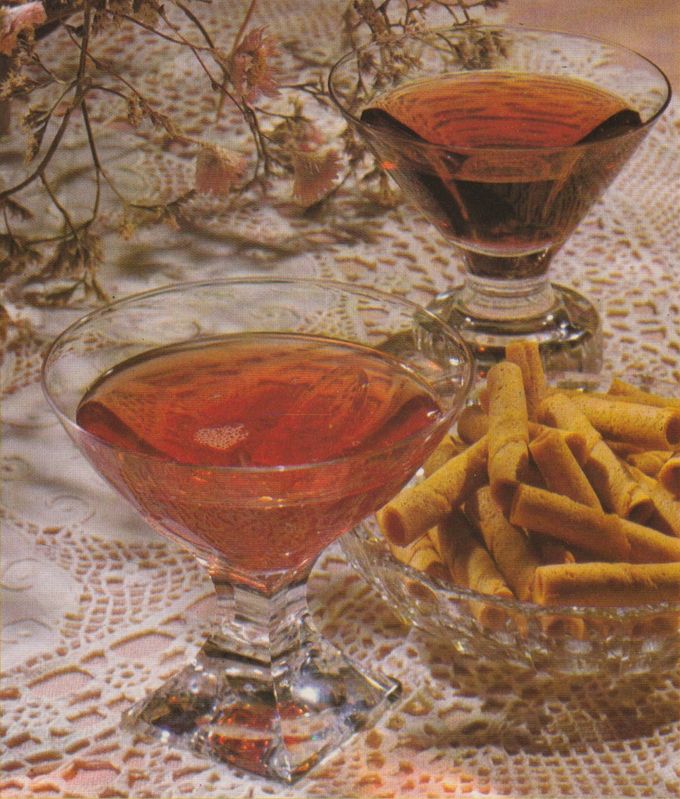 Zwei elegante Cocktailgläser mit bernsteinfarbener Flüssigkeit stehen auf einer gehäkelten Spitzendecke, daneben eine Glasschale mit goldbraunen gerollten Waffelröllchen. Getrocknete Blumen im Hintergrund runden die Dekoration im typischen Stil der 1970er-Jahre ab.