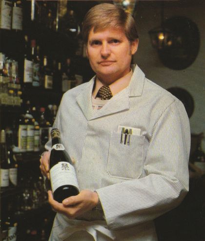 Ein Verkäufer in blau-weiß gestreifter Arbeitskleidung mit Krawatte präsentiert eine Weinflasche vor einem Regal voller Spirituosen und Weinflaschen.