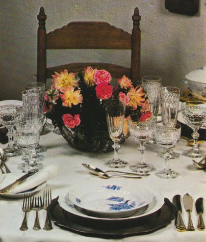 Ein festlich gedeckter Tisch mit weißer Tischdecke, Silberbesteck und geschliffenem Kristallglas, in der Mitte steht ein dunkler Blumenstrauß mit rosa und gelben Dahlien. Im Vordergrund liegen weiße Servietten mit blauem Blumenmuster auf den Tellern, dahinter ein antiker Holzstuhl.