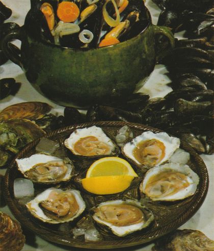 Sechs frische Austern auf der Halbschale sind auf einem Teller mit gedrehter Kordel-Dekoration arrangiert, in der Mitte liegt eine halbe Zitrone. Im Hintergrund steht eine grüne Suppenterrine gefüllt mit dampfenden Miesmuscheln, umgeben von weiteren Muschelschalen.