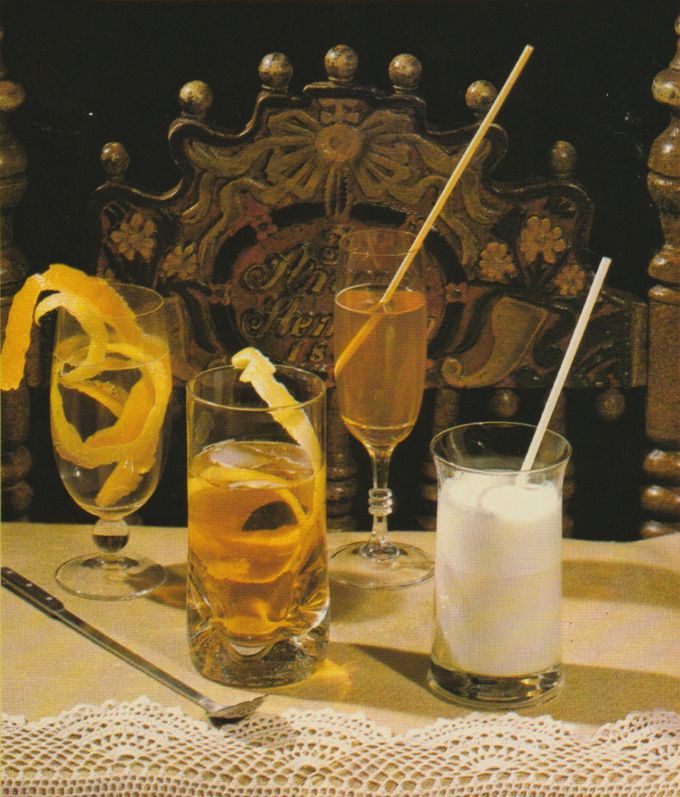 Vier verschiedene Cocktails in Gläsern vor einem verzierten Tisch. Von links: Ein Glas mit spiralförmig geschnittener Zitronenschale, ein Tumbler mit bernsteinfarbenem Drink und Zitronenspirale, ein Sektkelch mit Cocktail und Trinkhalm, und ein Tumbler mit milchigem Drink, Eiswürfeln und Trinkhalm. Im Vordergrund liegt ein Barlöffel.