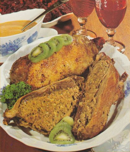 Knusprig gebratene Wildenten auf einer ovalen Servierplatte, garniert mit Kiwischeiben und Petersilie. Die Enten sind goldbraun gebraten und aufgeschnitten, sodass die Hackfleisch-Füllung sichtbar ist. Im Hintergrund stehen Weingläser mit Rotwein, eine Sauciere und Rotkohl.