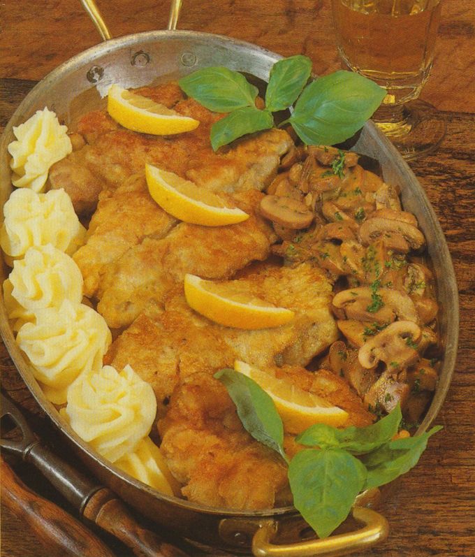 Ovale Servierplatte mit panierten Kalbsbries-Scheiben, garniert mit Zitronenscheiben und Basilikumblättern. Links am Rand dekorative Kartoffelpüree-Rosetten, rechts geschmorte Champignons mit Petersilie. Im Hintergrund ein Glas Weißwein auf Holztisch.