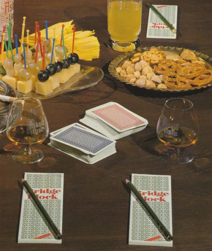 Ein gedeckter Spieltisch mit Käsewürfeln und Weintrauben auf Spießen, Salzgebäck auf einer Glasplatte, Schwenkern mit Cognac, Spielkarten und Bridge-Blöcken.