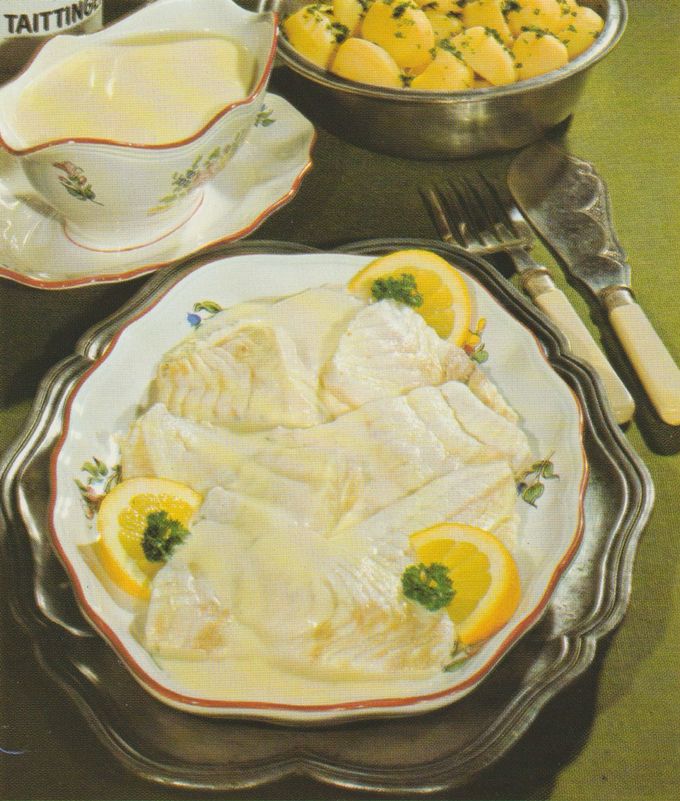 Pochierte Steinbuttfilets mit cremiger Sahnesauce auf einem Teller, garniert mit Zitronenscheiben und Petersilie. Im Hintergrund eine passende Sauciere und eine Schüssel mit Salzkartoffeln. Das Gedeck ist auf einer Tischdecke mit Serviette und Besteck angerichtet.