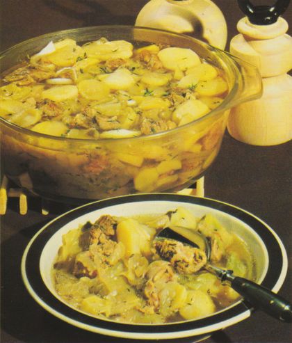 Ein Kartoffel-Hackfleisch-Auflauf in einer durchsichtigen Glasschüssel, daneben ein angerichteter Teller mit einer Portion des Gerichts und Servierbesteck. Im Hintergrund sind geschnittene Zwiebeln und ein gelber Salzstreuer zu sehen.