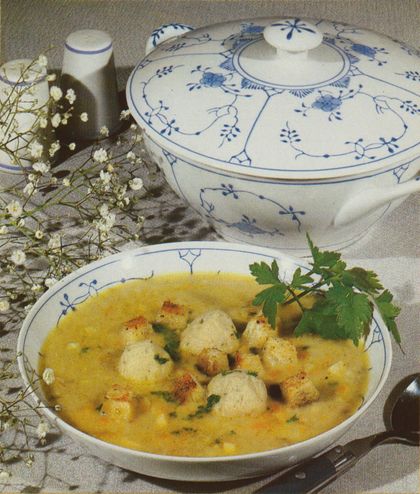 Eine cremige gelbe Suppe mit Semmelknödeln und frischer Petersilie, serviert in einer weißen Porzellanschüssel mit blauem Blumenmuster. Im Hintergrund steht eine passende Suppenterrine, daneben Schleierkraut als Dekoration auf beigem Leinen.