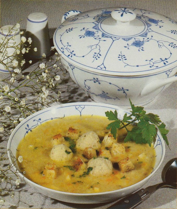 Eine cremige gelbe Suppe mit Semmelknödeln und frischer Petersilie, serviert in einer weißen Porzellanschüssel mit blauem Blumenmuster. Im Hintergrund steht eine passende Suppenterrine, daneben Schleierkraut als Dekoration auf beigem Leinen.