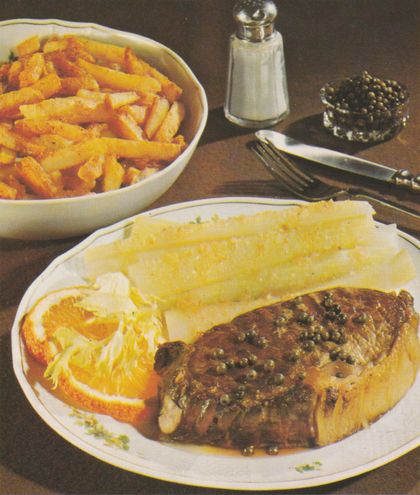 Gebratenes Rumpsteak mit grünem Pfeffer und Sauce auf einem Teller, garniert mit Orangenscheiben und Staudensellerie. Im Hintergrund eine Schale mit Pommes frites, Salzstreuer und eine kleine Kristallschale mit grünem Pfeffer. Besteck liegt neben dem Teller.