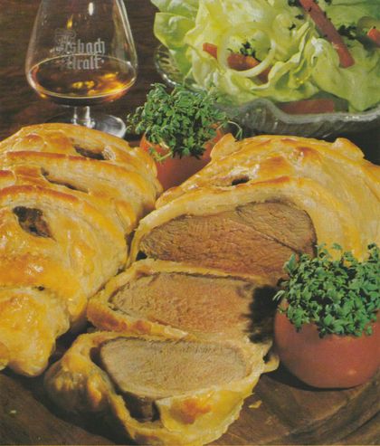 In Blätterteig gebackene Schweinefilets im Teigmantel, angeschnitten und aufgefächert, sodass das rosa-braune Fleisch sichtbar ist. Der goldbraun gebackene Teig ist mit aufgeklebten Teig-Ornamenten verziert. Als Garnitur dienen mit frischer Kresse gefüllte Ton-Töpfchen sowie halbierte, ausgeholte Tomaten mit Kresse. Im Hintergrund ein Schwenker mit Weinbrand sowie ein Teller mit Kopfsalat.