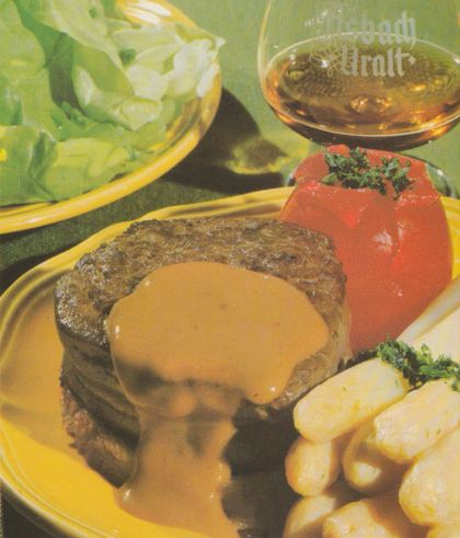 Gebratenes Rinderfiletsteak mit hellbrauner Soße auf gelbem Teller, dazu gefüllte Tomate mit Kräutern und Salzkartoffeln mit Petersilie. Im Hintergrund eine Schale mit grünem Salat und ein Glas Asbach Uralt.