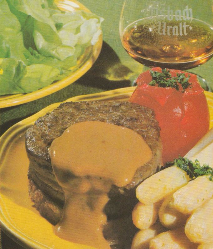 Gebratenes Rinderfiletsteak mit hellbrauner Soße auf gelbem Teller, dazu gefüllte Tomate mit Kräutern und Salzkartoffeln mit Petersilie. Im Hintergrund eine Schale mit grünem Salat und ein Glas Asbach Uralt.