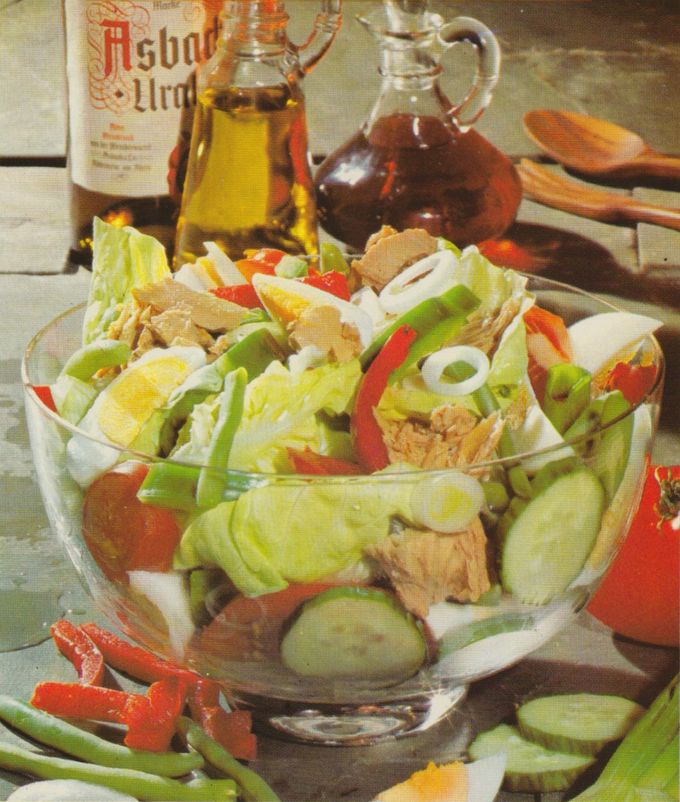 Eine große Glasschüssel mit buntem Salat aus Eisbergsalat, Gurkenscheiben, Tomaten, grünen und roten Paprikastreifen, Zwiebelringen und Thunfischstücken. Im Hintergrund stehen Flaschen mit Öl und Essig sowie Holzlöffel auf einem weißen Holztisch.