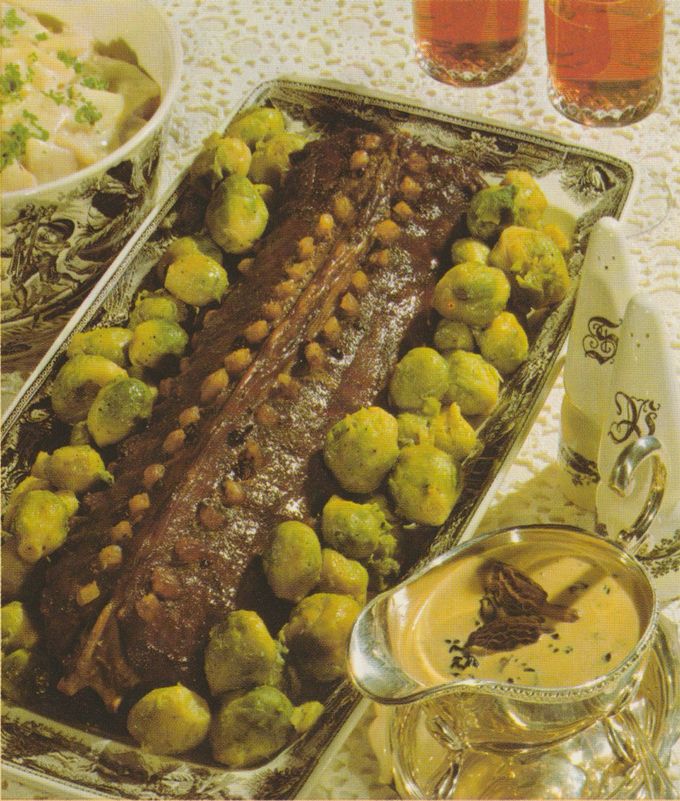 Gebratener Rehrücken auf einer verzierten Silberplatte, umgeben von buttrig glänzenden Rosenkohl-Röschen und garniert mit Morcheln. Rechts unten eine Sauciere mit heller Pilzsauce, darauf MOrcheln. Im Hintergrund eine Schüssel mit Sellerie, zwei Gläser Asbach Uralt und Salz- und Pfefferstreuer auf strukturierter Tischdecke.