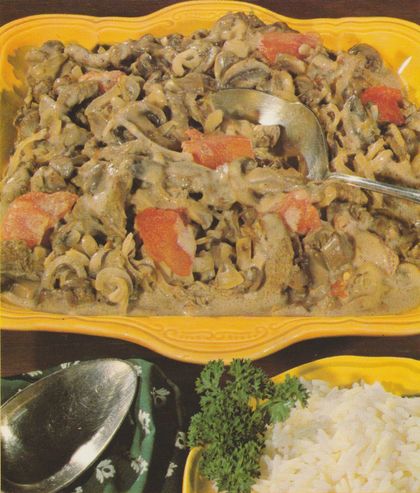 Spiralnudeln mit Rindfleischstreifen in heller Sahnesauce, durchsetzt mit Champignons und leuchtend roten Tomatenstücken, serviert auf einer gelben Platte mit Löffel. Im Vordergrund frische Petersilie und weißer Reis in einer gelben Schale.