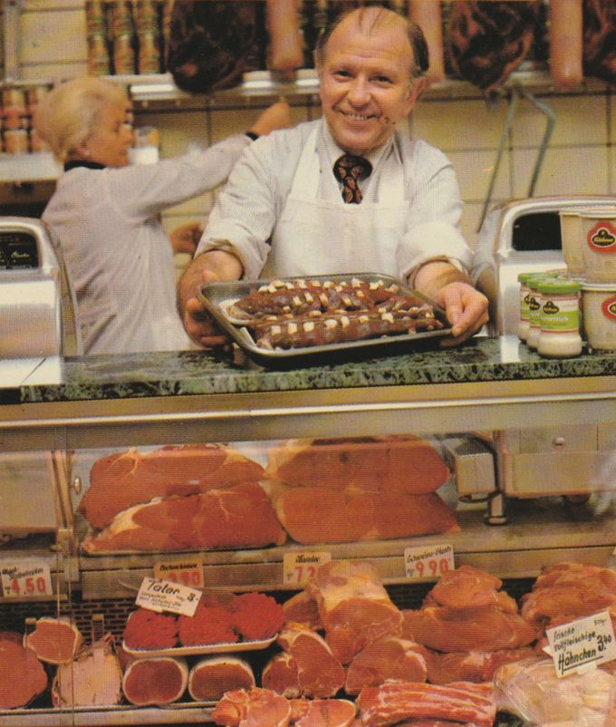 Ein lächelnder Metzger in weißer Schürze präsentiert in einer Fleischerei ein Tablett mit glasierten Bratwürsten, die mit Zwiebelringen garniert sind. In der Auslage davor liegen verschiedene Schinken- und Fleischsorten mit Preisschildern.