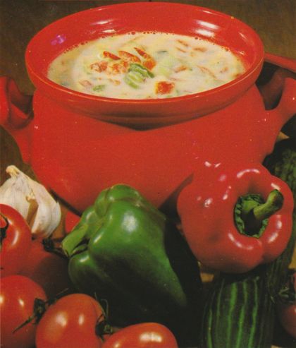 Eine cremige helle Suppe in einem orangefarbenen Topf mit Henkeln, garniert mit Speckwürfeln und Lauchringen. Drumherum sind frische Zutaten arrangiert: rote und grüne Paprika, Knoblauch und Tomaten.