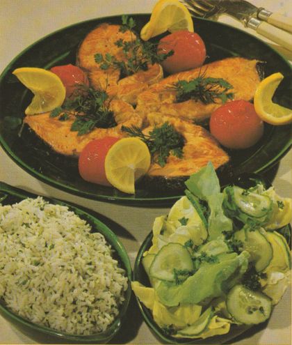 Gebratene Lachsscheiben auf dunklem Teller, garniert mit Petersilie, Dill, gegrillten Tomaten und Zitronenscheiben. Als Beilagen Kräuterreis und Kopfsalat mit Gurkenscheiben auf separaten Tellern.