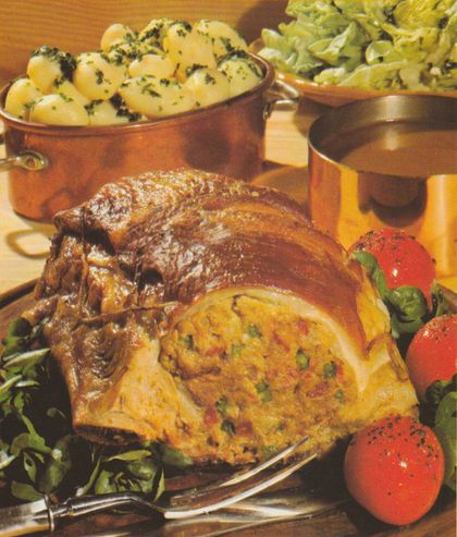 Gefüllte Kalbsbrust auf einem Bett aus grünem Salat, angeschnitten und mit einer gewürzten Hackfleisch-Füllung mit roten Pimiento-Streifen. Die Kruste ist goldbraun und glänzend. Im Hintergrund Kupfertöpfe mit Petersilienkartoffeln und Kopfsalat sowie eine Saucière. Garnitur aus Tomaten und frischem Grün.