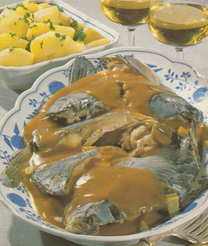 Mehrere Karpfenstücke in dickflüssiger brauner Sauce auf einem Teller mit blauem Blumenmuster. Im Hintergrund eine Schüssel mit petersilienbestreuten Salzkartoffeln und zwei Glas Weißwein.