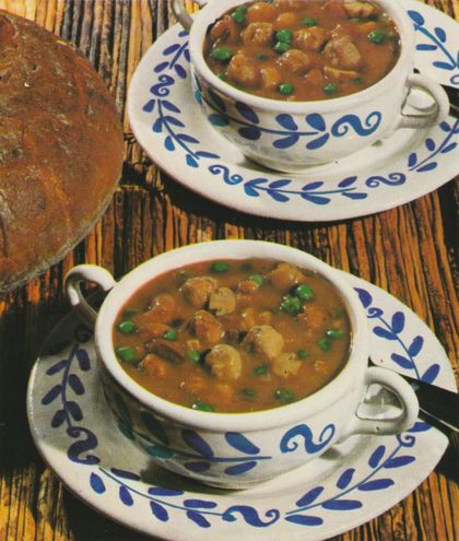 Zwei Suppentassen mit blauem Blattmuster sind gefüllt mit einer braunen Suppe, in der Champignons, grüne Erbsen und Fleischstücke schwimmen. Daneben liegt ein Laib dunkles Brot auf einer Holzoberfläche.