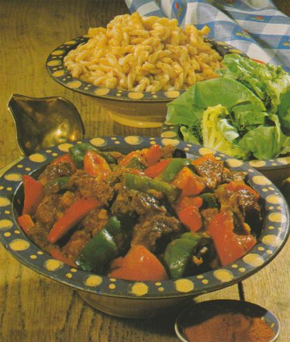Paprika-Schweinegulasch mit roten und grünen Paprikastreifen in dunkler Sauce, serviert in einer schwarz-gelb gepunkteten Schüssel. Im Hintergrund eine gleichartige Schüssel mit gedrehten Nudeln sowie frischer Kopfsalat als Beilage. Auf einem Holztisch angerichtet, im Hintergrund ein blau-weiß kariertes Tischtuch.