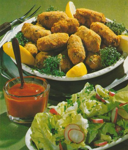 Panierte Geflügel–Kroketten auf einer ovalen Platte, garniert mit Petersilie und Zitronenscheiben. Daneben ein Glas Tomatensauce mit Löffel und eine Schüssel gemischter Blattsalat mit Radieschen.
