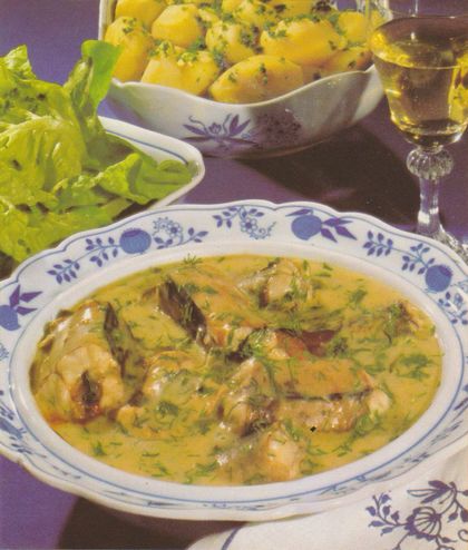 Aalstücke in cremiger Dillsauce auf einem gemusterten Teller, serviert mit Pellkartoffeln in einer Schale und einem Glas Weißwein. Im Hintergrund Blattsalat als Beilage.