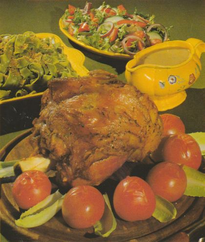 Gebratene Kalbshaxe auf einer Servierplatte, umgeben von Kirschtomaten und Salatblättern. Im Hintergrund eine gelbe Sauciere mit Blumenmuster sowie zwei Salatschüsseln – eine mit grünen Bandnudeln und gerösteten Semmelbröseln, die andere mit gemischtem Salat aus Tomaten, Zwiebeln und Pilzen.