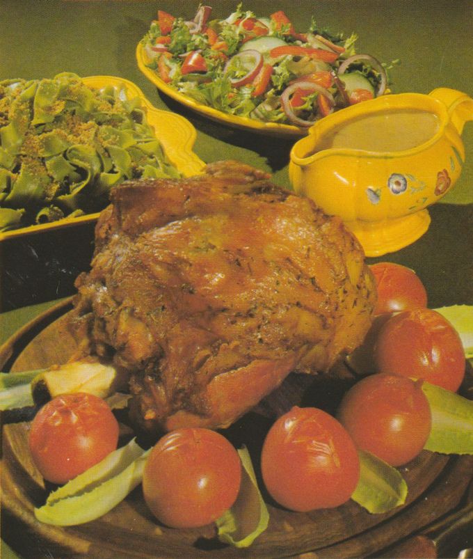 Gebratene Kalbshaxe auf einer Servierplatte, umgeben von Kirschtomaten und Salatblättern. Im Hintergrund eine gelbe Sauciere mit Blumenmuster sowie zwei Salatschüsseln – eine mit grünen Bandnudeln und gerösteten Semmelbröseln, die andere mit gemischtem Salat aus Tomaten, Zwiebeln und Pilzen.