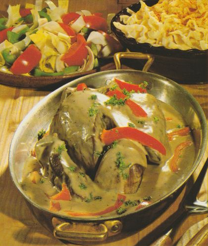 Kupferfarbene Kasserolle mit geschmortem Kalbsherz in heller, sämiger Sauce, garniert mit roten Paprikastreifen, Sahnetupfern und Petersilie. Im Hintergrund Holzschale mit Chicoréesalat und Tomaten sowie eine Schale mit Butterspätzle.