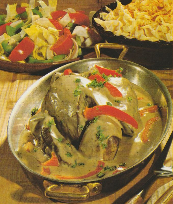 Kupferfarbene Kasserolle mit geschmortem Kalbsherz in heller, sämiger Sauce, garniert mit roten Paprikastreifen, Sahnetupfern und Petersilie. Im Hintergrund Holzschale mit Chicoréesalat und Tomaten sowie eine Schale mit Butterspätzle.